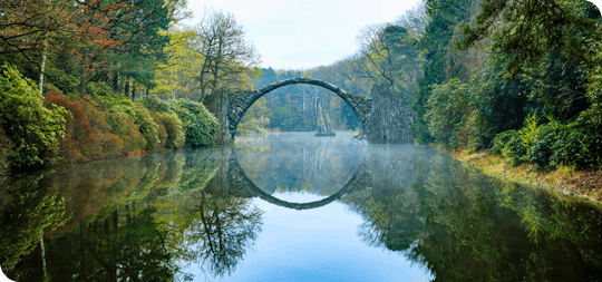 Image of old stone bridge crossing a lake with forests all around, the reflection makes the bridge look like a circle.