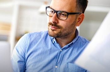 A man wearing a blue checkered shirt holds a stack of papers, seated in a modern office environment with a laptop in front of him