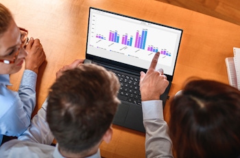 Three people analyse graphs displayed on a laptop during a meeting, with notebooks and a smartphone on the table