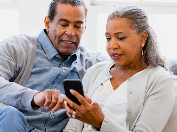Older couple sitting on couch, woman holding smartphone while man points at the screen, both focused on the device