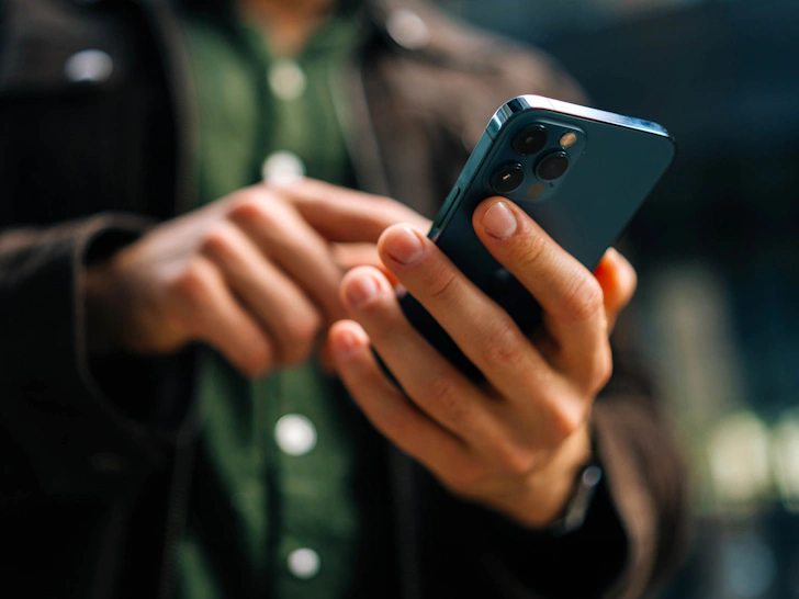 Person wearing a dark jacket and green shirt using a blue smartphone with a triple camera setup in an outdoor setting