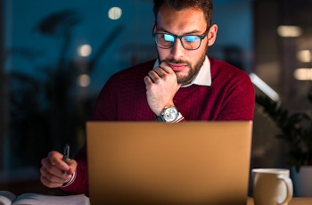 A person in a red sweater sits at a desk, contemplating with a pen in hand and a coffee mug beside a laptop, illuminated by soft light