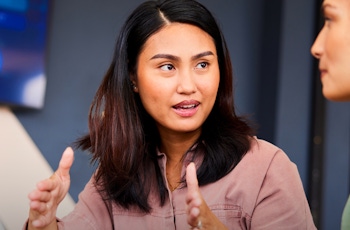 Two women engaged in a discussion in a modern office setting, with a laptop and a presentation screen in the background