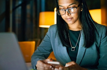A woman in business attire sits at a desk, using a smartphone while focused on a laptop screen, illuminated by warm lamp light