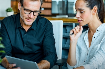 Two people engaged in a discussion at a desk, one holding a tablet while the other ponders, with greenery in the foreground