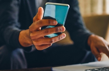 A person in a suit holds a smartphone in one hand while typing on a laptop with the other, seated on a couch. Soft lighting in background