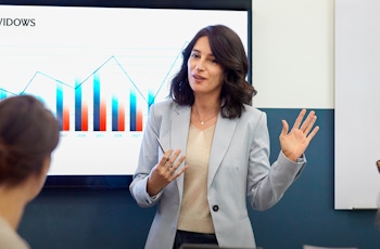 Young female with long brown hair and wearing a light grey jacket presents in front of four seated work colleagues whilst standing in front of a screen with a blue and red data chart beside a scottish widows logo