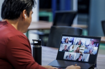 A person wearing earbuds is attending a video conference on a laptop, displaying multiple participants on the screen in a workspace setting