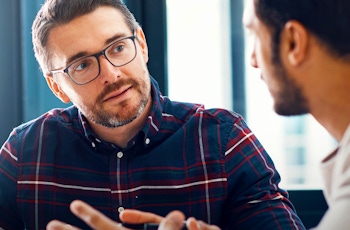 Two men engaged in a discussion at a table, surrounded by bright windows and a laptop, illustrating a collaborative work atmosphere