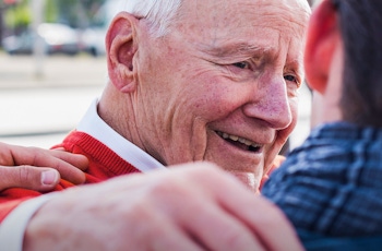 An older man in a red sweater rests comfortably, while another person gently places a hand on his shoulder, showing support and warmth