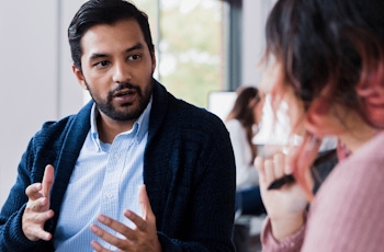 A person in a blue cardigan gestures while engaged in conversation with a woman, amidst a bright, modern office setting