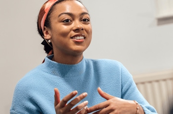 A person wearing a blue sweater gestures while speaking in a discussion setting, with a play button overlay on the image