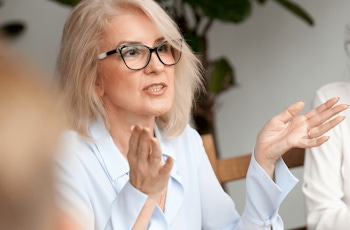 A woman in a light blue blouse gestures while speaking during a meeting, surrounded by greenery and participants in a bright setting