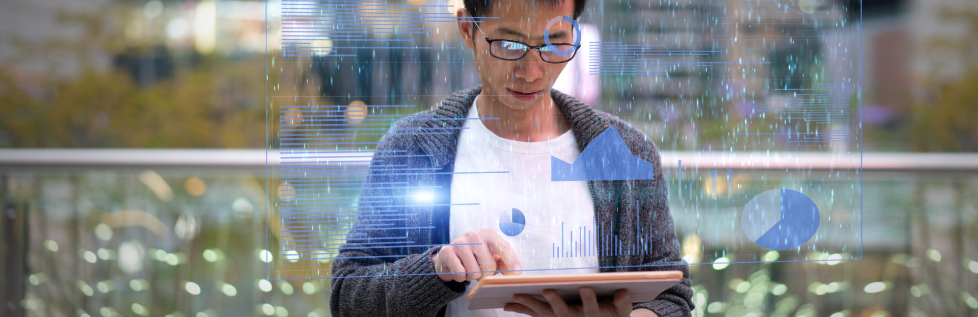 Man with a grey jumper and dark rimmed glasses working on a tablet with an overlay of blue coloured charts and graphs