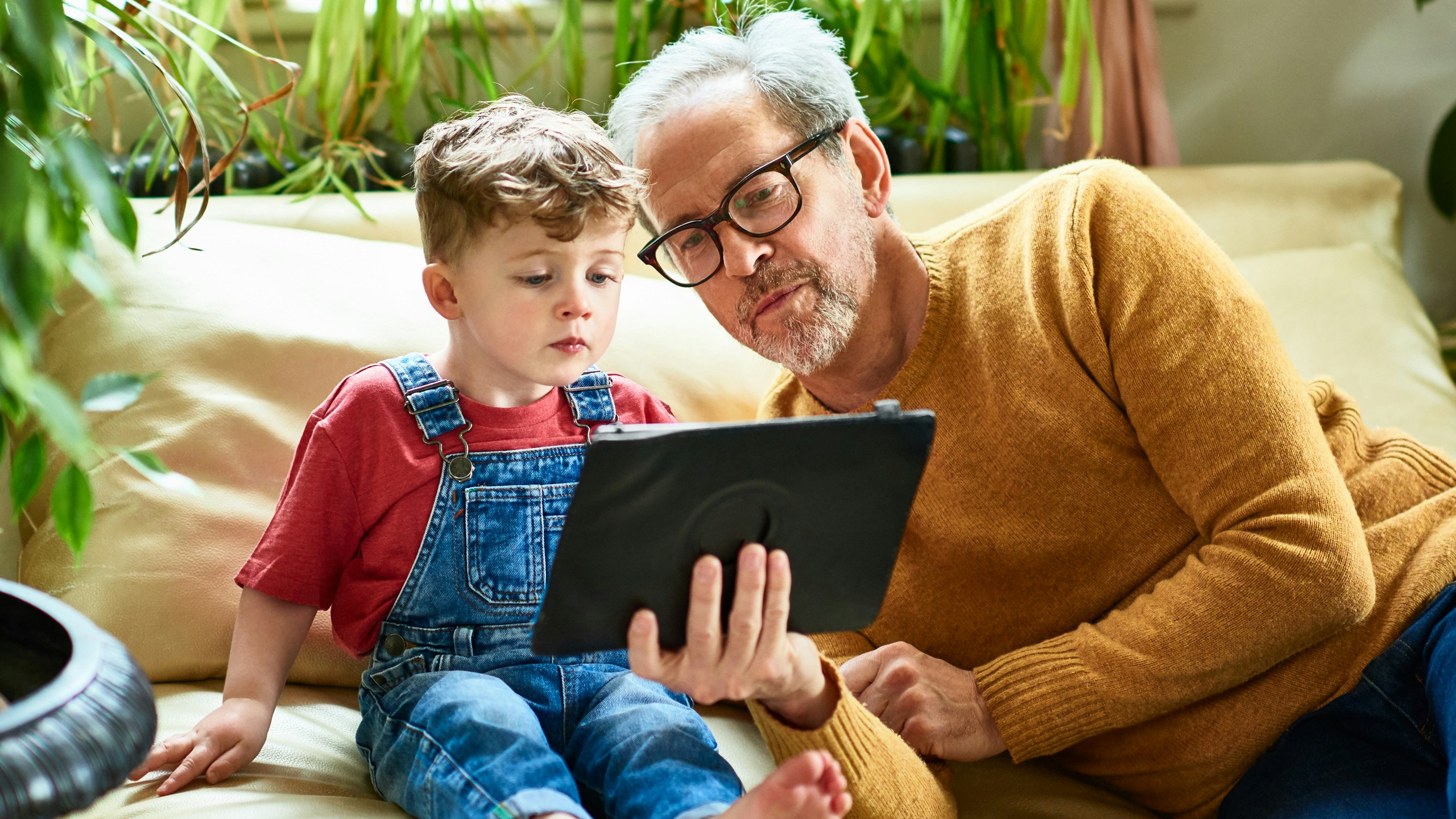 Image of grandad with yellow jumper sitting showing his young grandson something on a tablet device