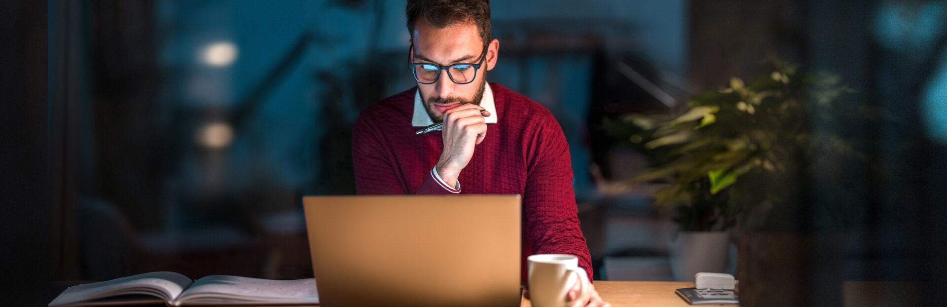 Young male with beard and glasses, wearing a maroon jumper sits at a desk in a dark office late at night, working on a laptop whilst holding a cup of coffee