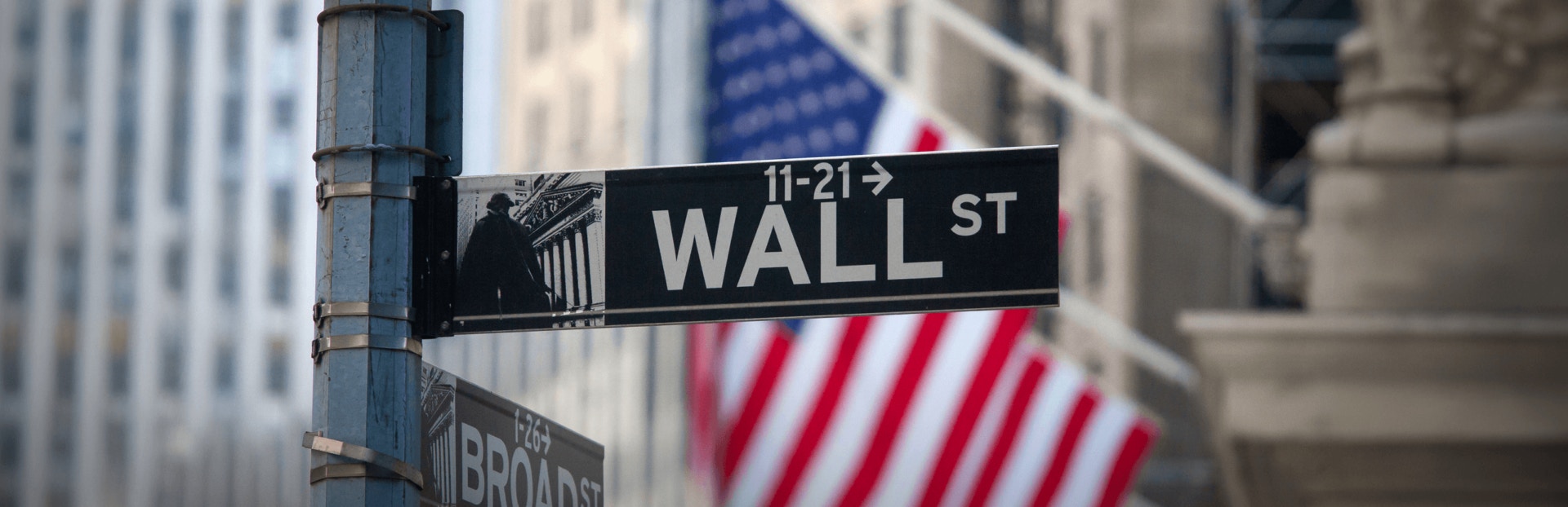 Street signs for Wall Street and Broad Street, with a blurred American flag in the background