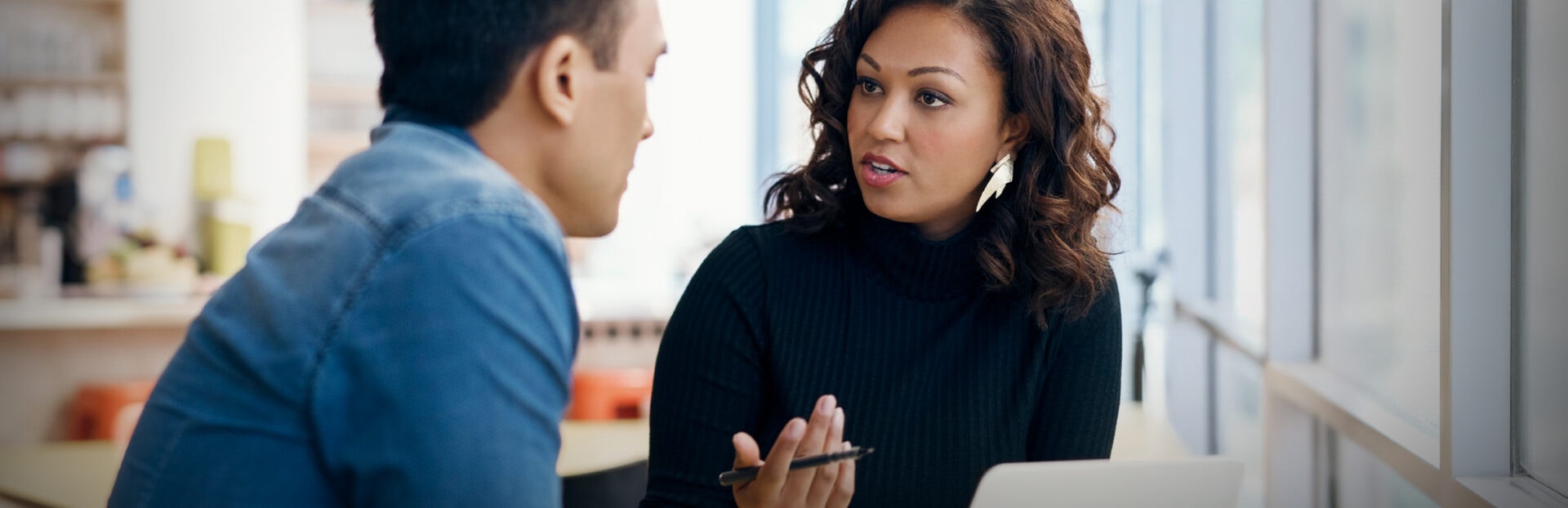 Two people have a discussion at a table in a bright office, one gesturing while holding a pen, with a laptop visible in front of them