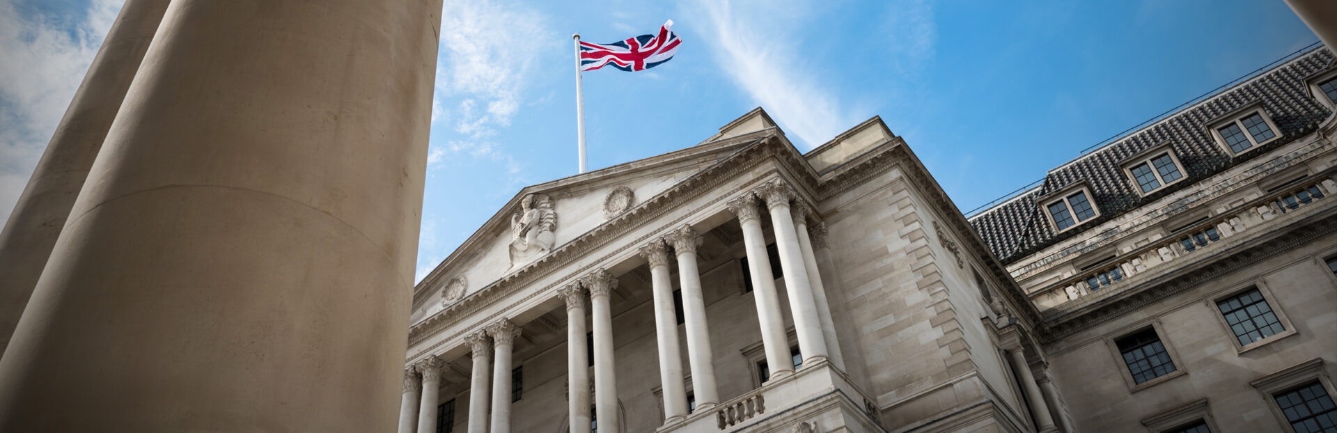 The Bank of England's façade with British flag flying, framed by tall columns against a clear blue sky