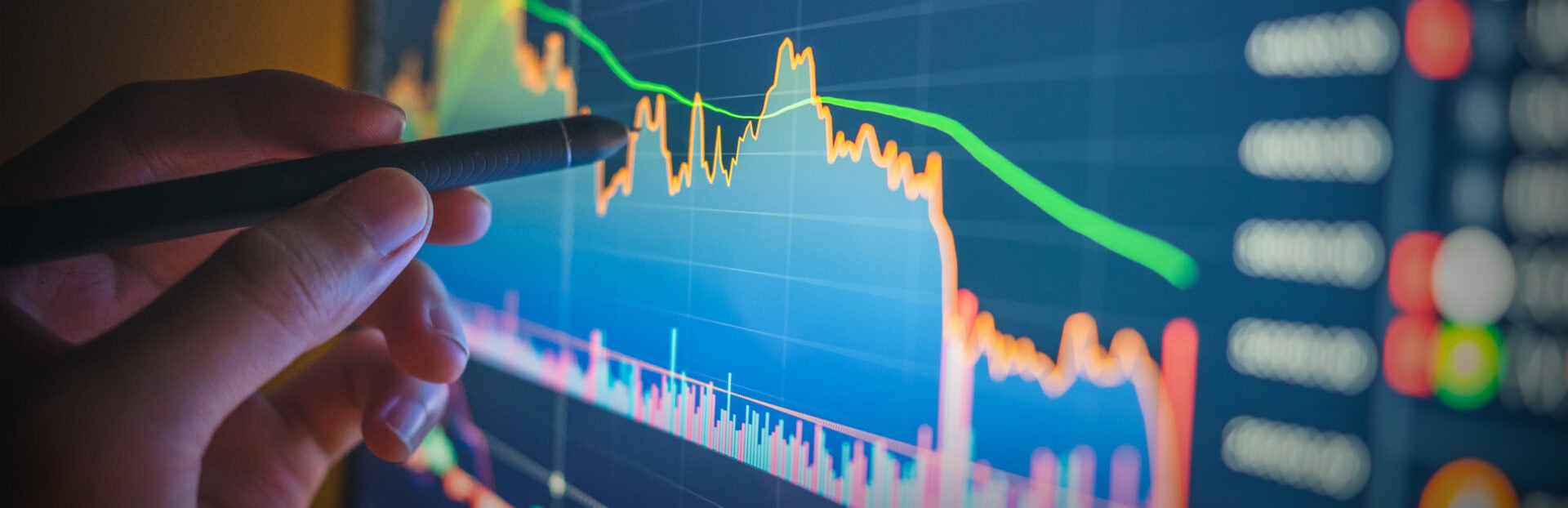 A hand holding a pen points at a vibrant financial chart on a screen displaying fluctuating prices and trends in various colors