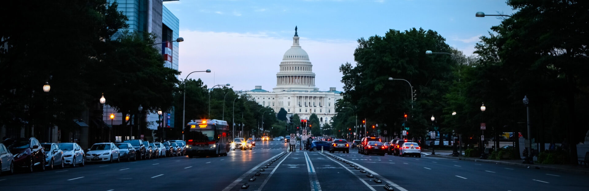 Dusk view of the U.S. Capitol building in Washington DC, framed by busy streets and traffic lights, with cars and a bus in the foreground