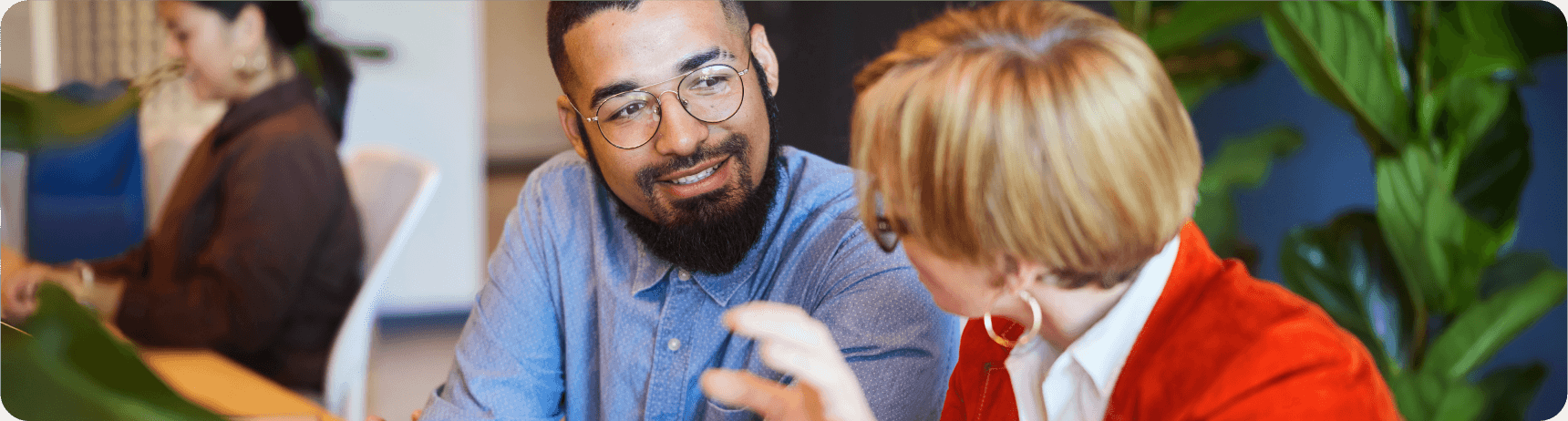 vc-additional-resources-01 A woman with short blonde hair and a red cardigan engages in conversation with a man in a blue shirt, surrounded by green plants
