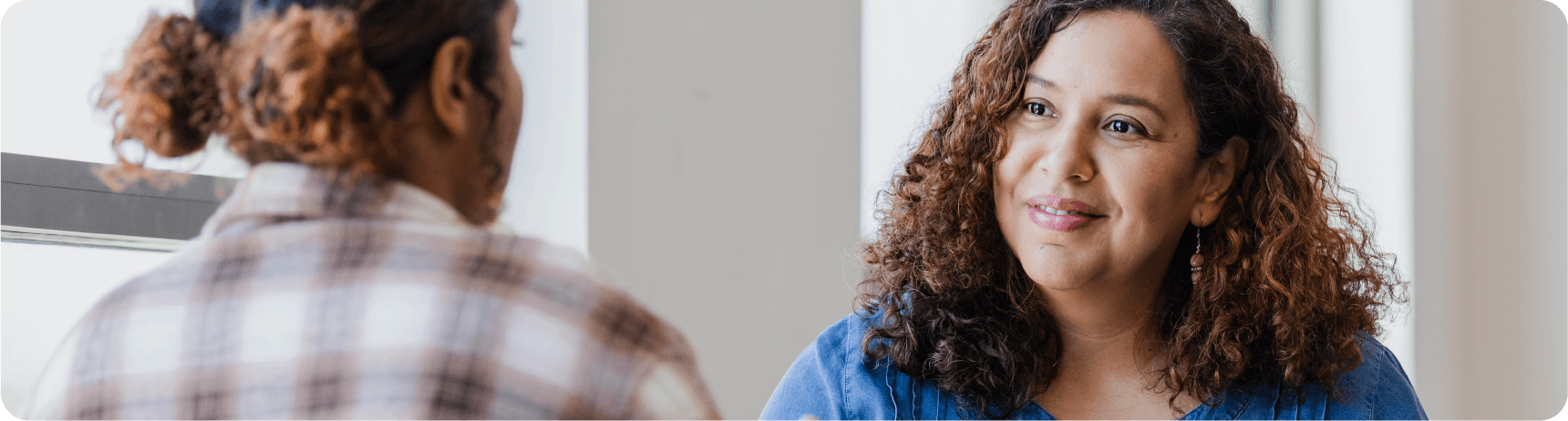 vc-additional-resources-04 Two women are engaged in a conversation indoors, one wearing a plaid shirt and the other in a blue top, in a well-lit setting